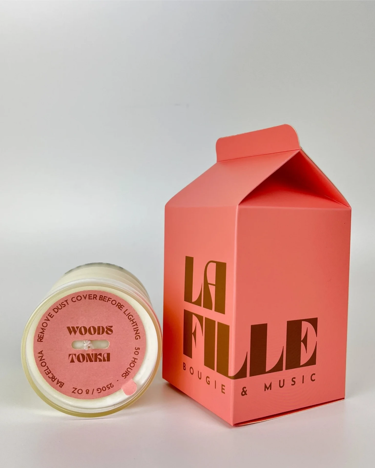 WOODS & TONKA scented candle in a glass vessel beside its rose and chocolate La Fille Milk Box packaging.