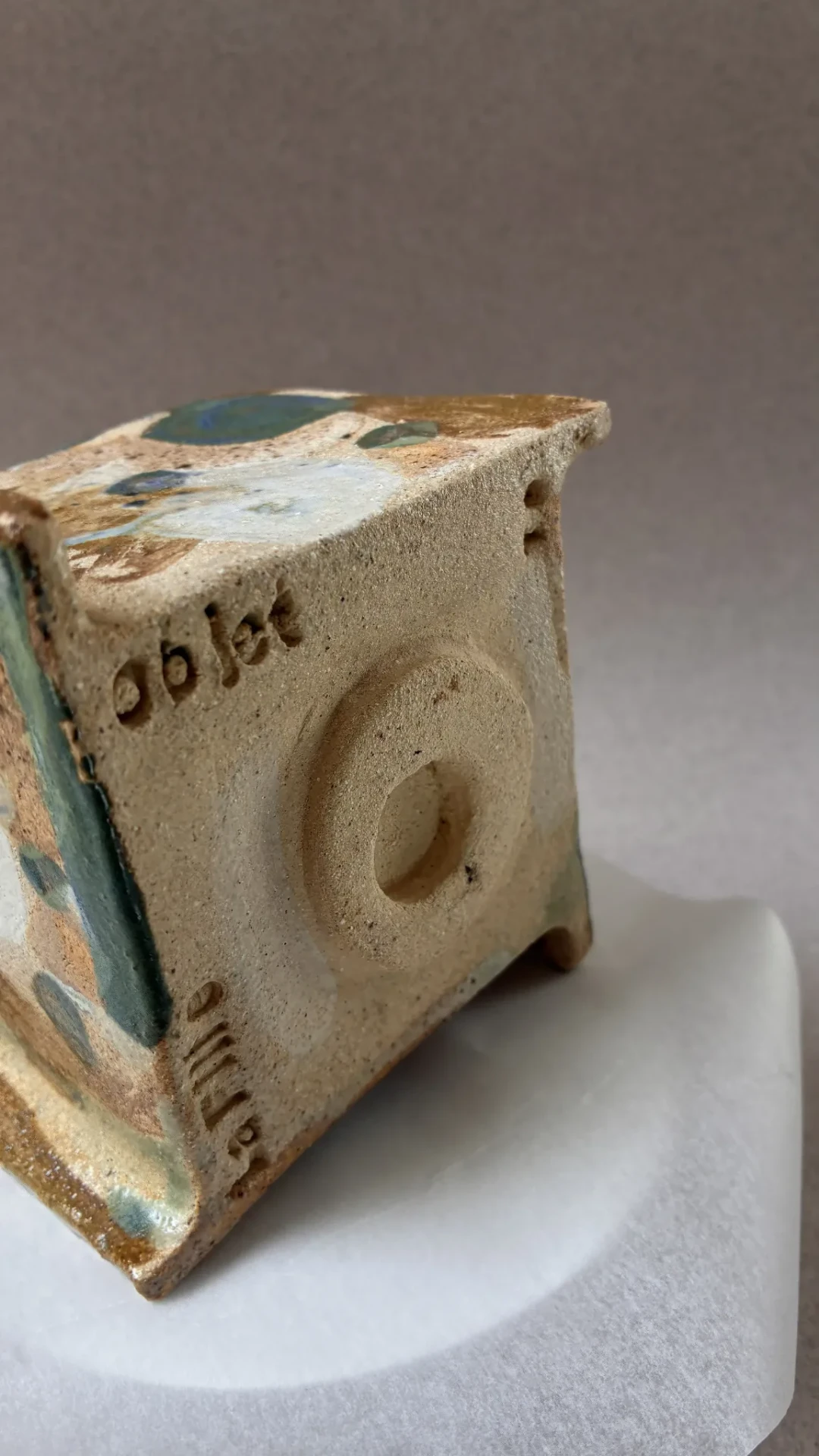 Handmade stamped stoneware object with green and blue glaze on white marble.