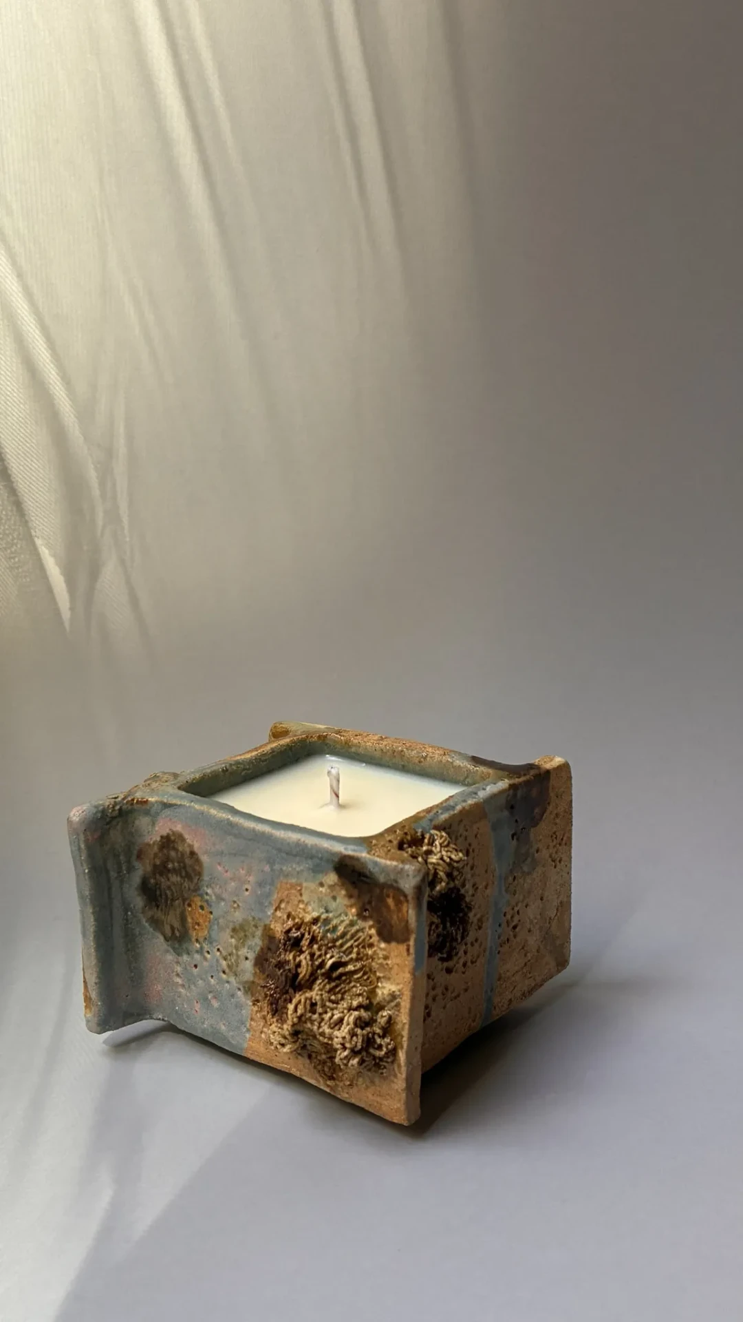 Textured ceramic candle in soft light beneath a pale curtain.