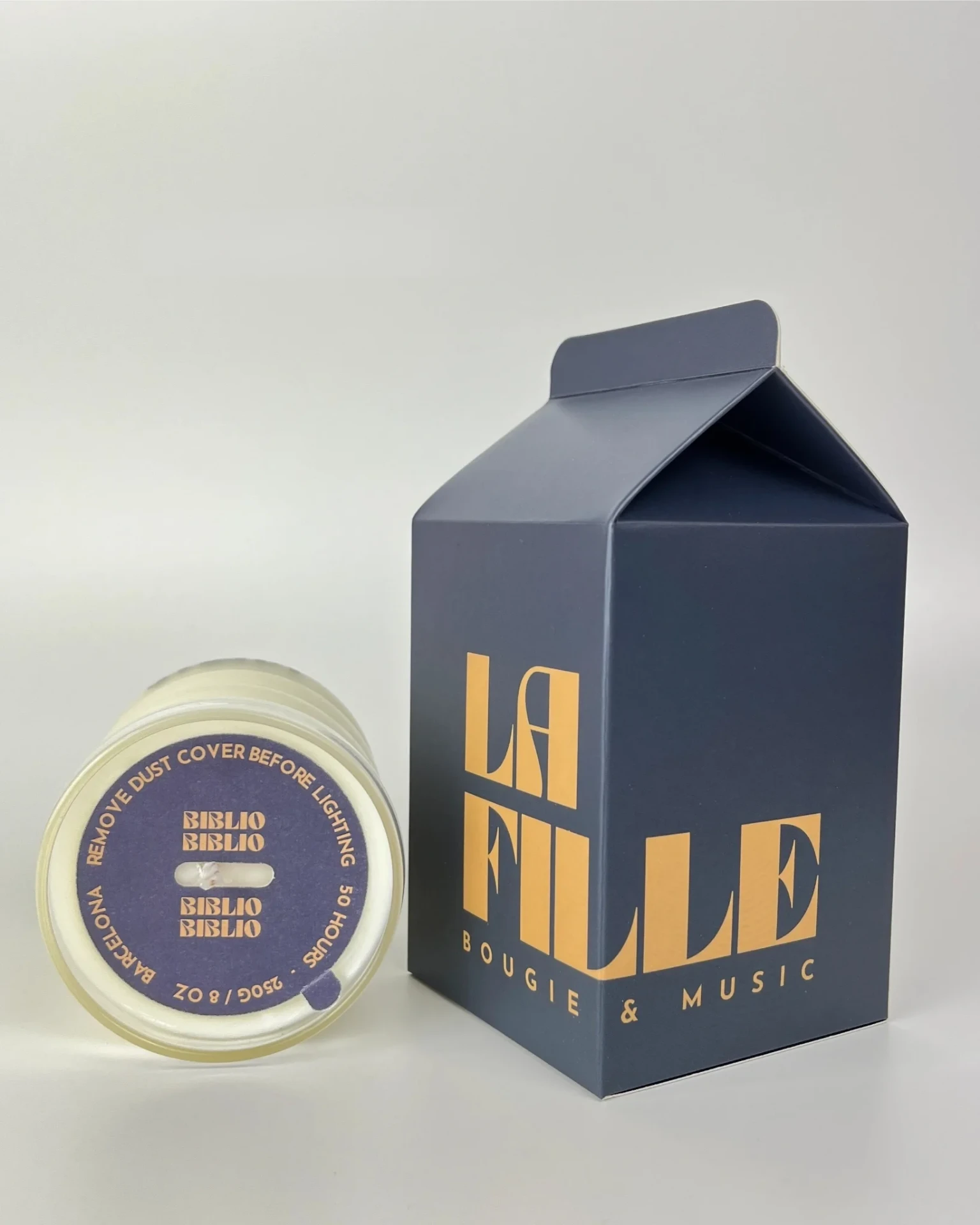 BIBLIOTHÈQUE scented candle in a glass vessel beside its navy blue and ochre La Fille Milk Box packaging.