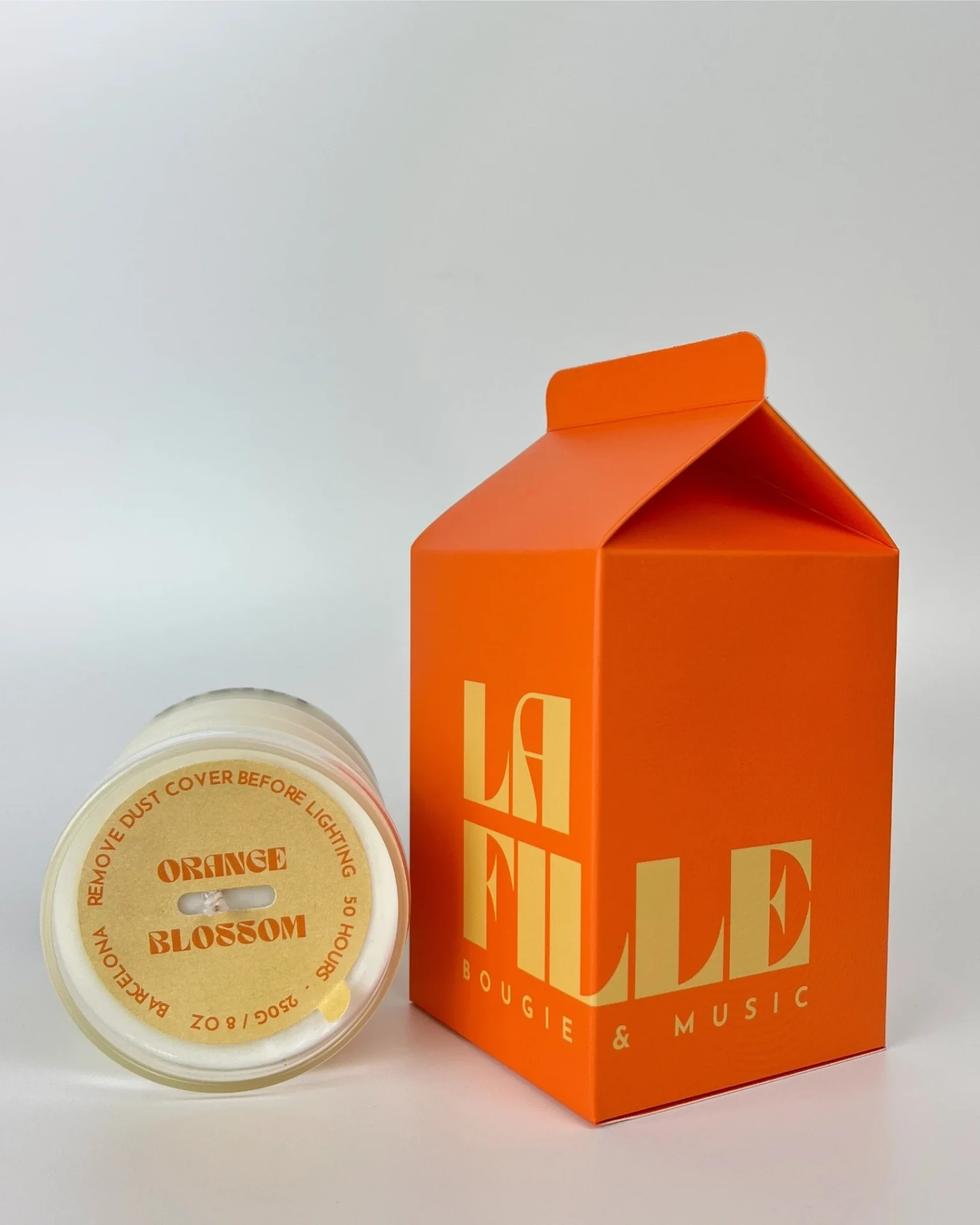 ORANGE BLOSSOM scented candle in a glass vessel beside its orange and vanilla La Fille Milk Box packaging.