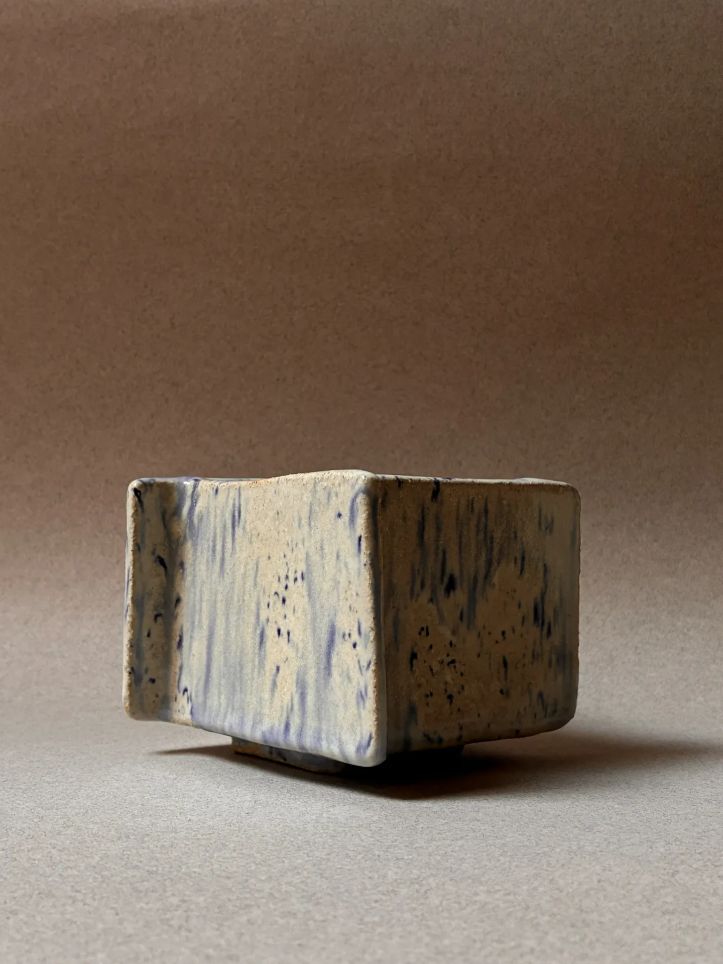 Square stoneware vessel with streaked blue glaze on a warm brown backdrop.
